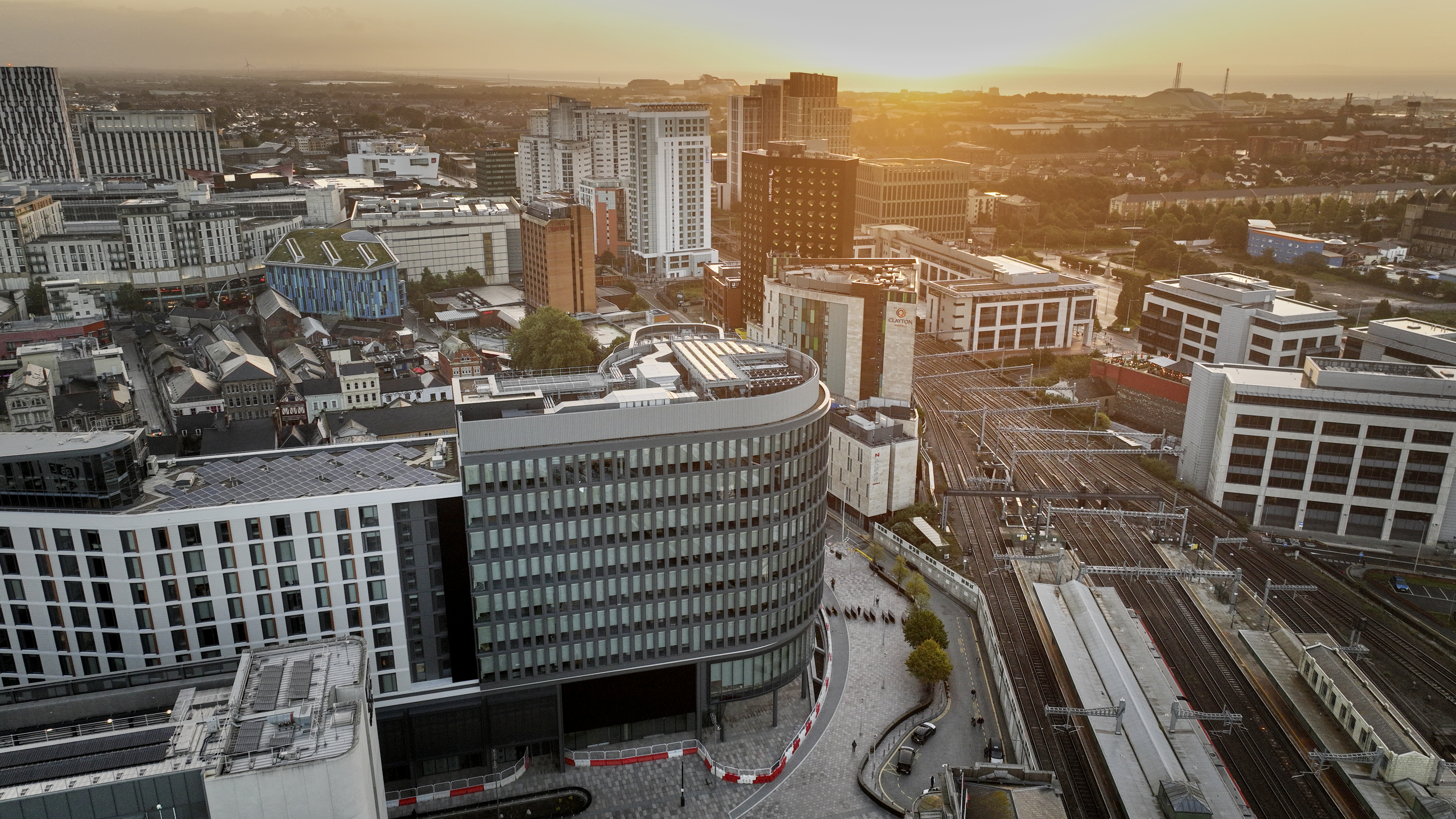 Aerial view of Cardiff Central Square at sunset. In the foreground is L&G's distinctive curved, multi-story office building with large windows. Surrounding are roads and railway tracks. The setting sun casts a warm golden glow over the city.