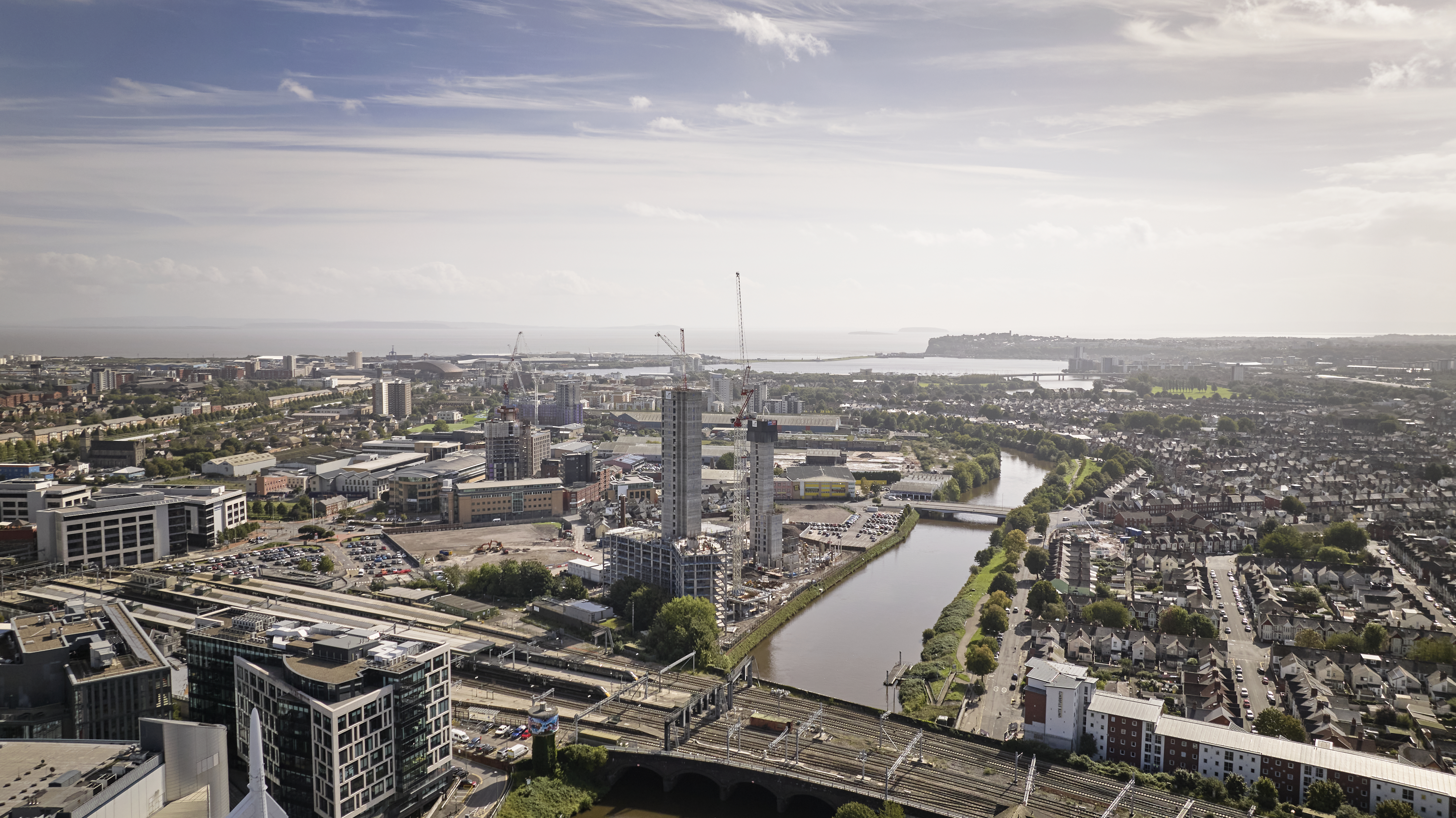 Aerial view of Cardiff showing multiple construction sites, cranes, and buildings in progress. The River Taff runs through the centre with bridges crossing it, surrounded by residential and commercial areas, roads, and railway tracks.