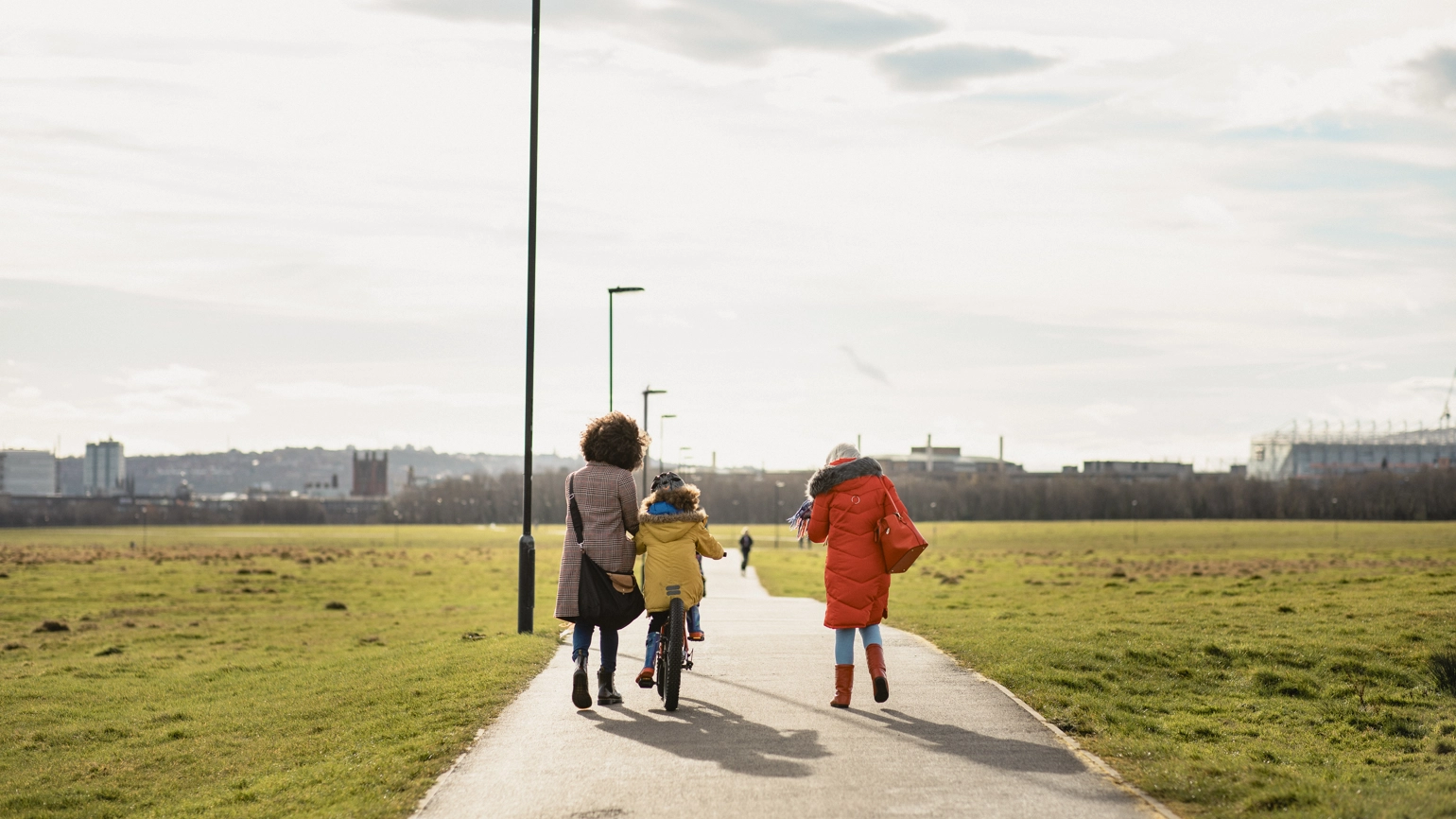 A wide paved path runs through a large green park under a bright, partly cloudy sky. Three people are walking away from the camera along the path: one person is walking with a child riding a bicycle, and another person is walking beside them. They are all wearing colourful winter coats. Tall streetlights line the path, and in the distance, there is a city skyline with low-rise buildings and a large stadium-like structure on the right. A wide paved path runs through a large green park under a bright, partly cloudy sky. Three people are walking away from the camera along the path: one person is walking with a child riding a bicycle, and another person is walking beside them. They are all wearing colourful winter coats. Tall streetlights line the path, and in the distance, there is a city skyline with low-rise buildings and a large stadium-like structure on the right.