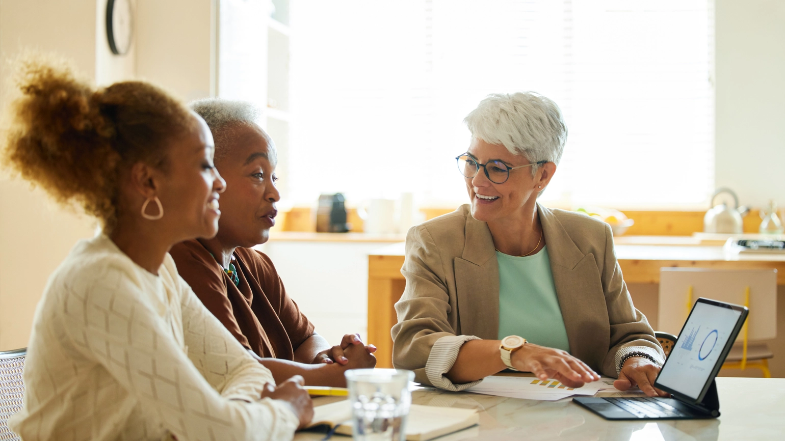 Three people seated at a table in a modern, in a well-lit room, engaged in discussion. A tablet is placed on the table in front of one person, along with documents and glasses of water. The background features a sofa and framed artwork of the walls. Three people seated at a table in a modern, in a well-lit room, engaged in discussion. A tablet is placed on the table in front of one person, along with documents and glasses of water. The background features a sofa and framed artwork of the walls.
