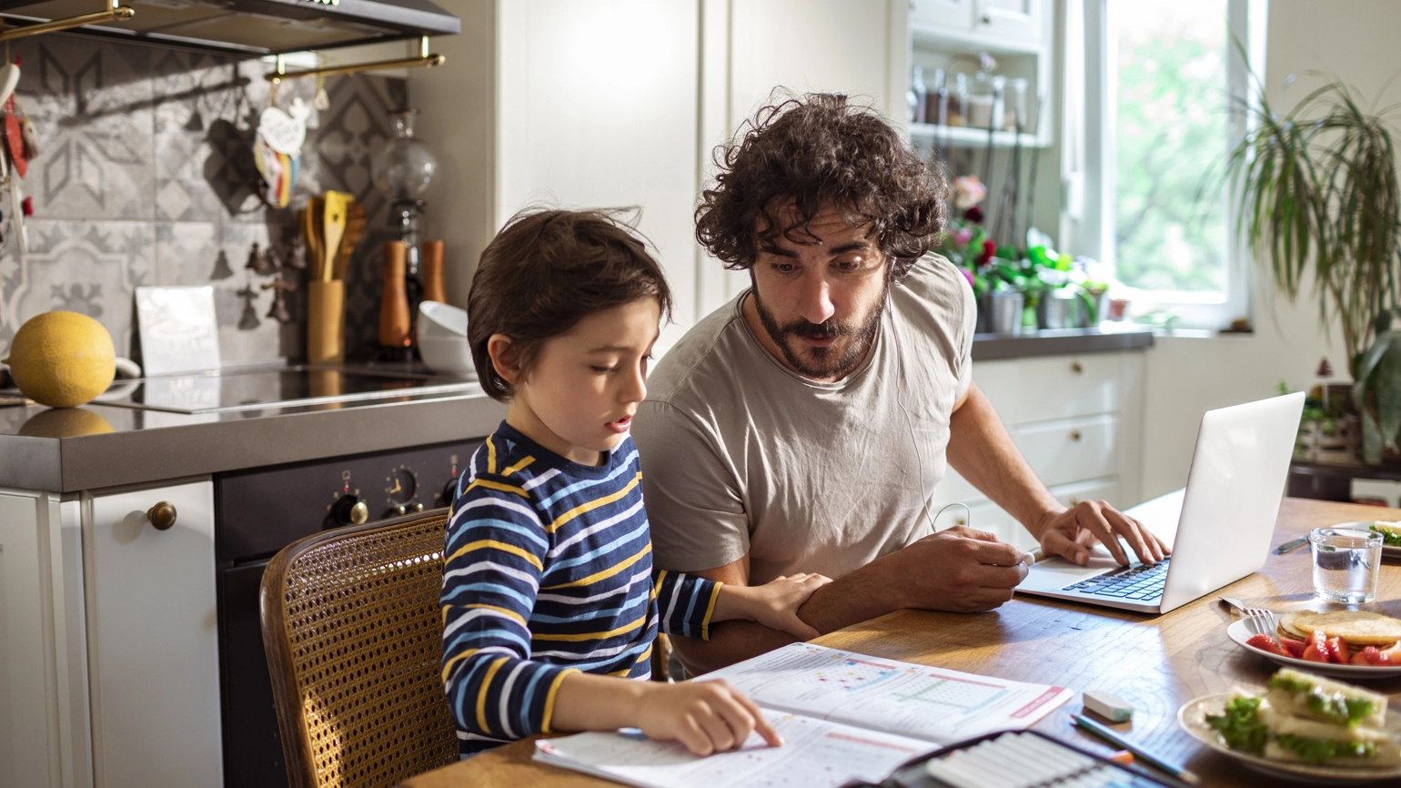 A man and a boy sit together in a kitchen, focused on a laptop as they work together on a project or homework. A man and a boy sit together in a kitchen, focused on a laptop as they work together on a project or homework.