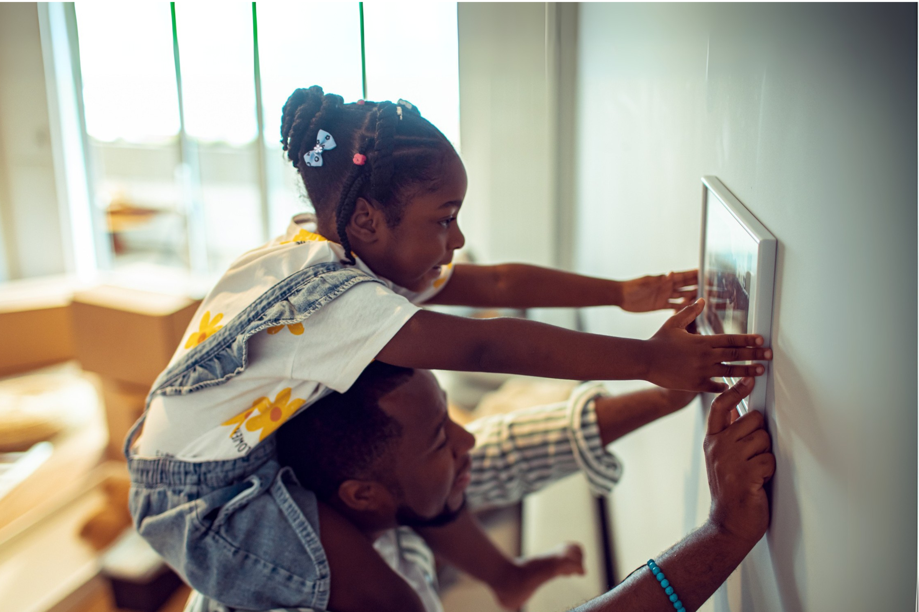 A child sits on an adult’s shoulders as they hang a picture frame on the wall of their new home.
