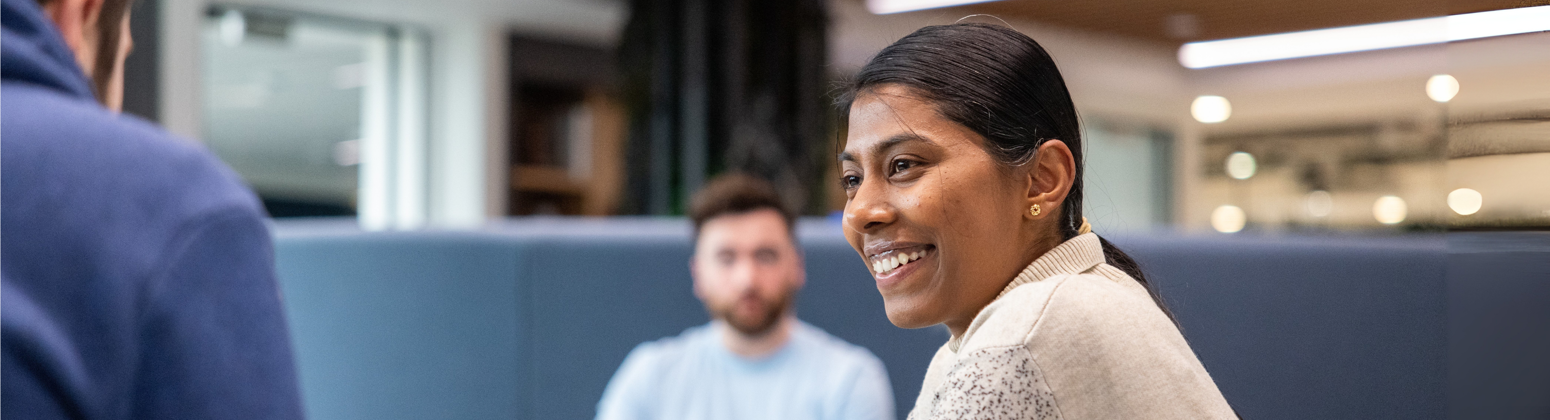 Two colleagues are seated around a table in a well-lit room with wooden ceiling panels. One wears a white and blue speckled jumper, another a navy blue hoodie., Two colleagues are seated around a table in a well-lit room with wooden ceiling panels. One wears a white and blue speckled jumper, another a navy blue hoodie.,