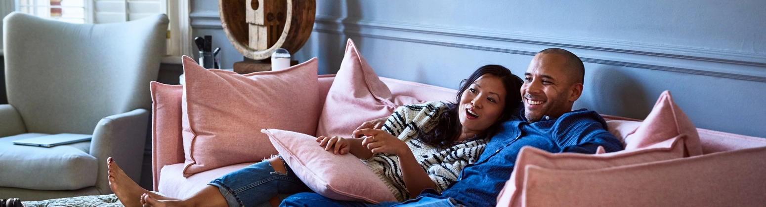 A man with his arm around a woman, sitting with their feet up on a pink sofa, smiling. A man with his arm around a woman, sitting with their feet up on a pink sofa, smiling.