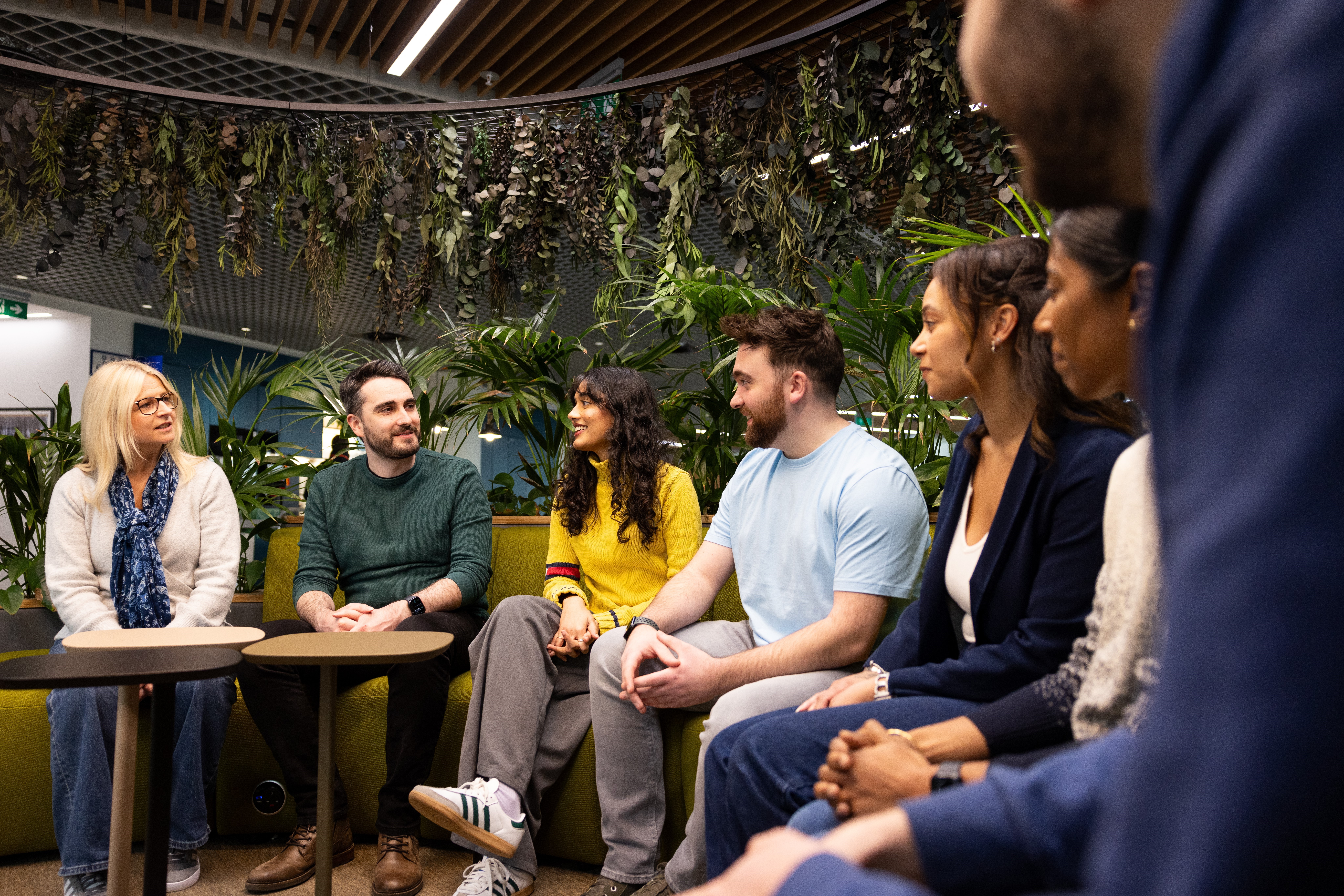 A group of people are seated in a semi-circle on green couches in Legal & General’s Cardiff office. The space is adorned with plants hanging from the ceiling.