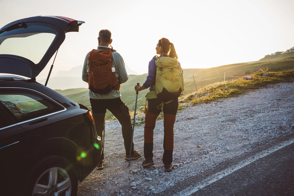 Two hikers standing beside an open car trunk on a gravel roadside, wearing backpacks and outdoor gear, looking toward sunlit grassy slopes and mountain peaks in the distance. Two hikers standing beside an open car trunk on a gravel roadside, wearing backpacks and outdoor gear, looking toward sunlit grassy slopes and mountain peaks in the distance.