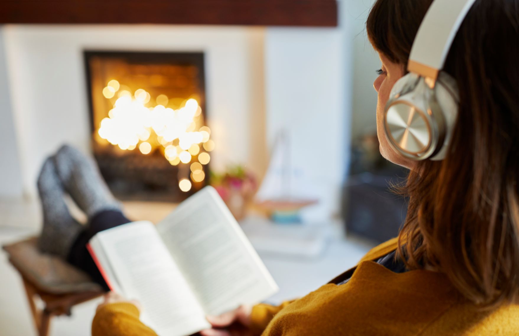 Female reading her book and listening to the music through headset in front of fire place in their living room