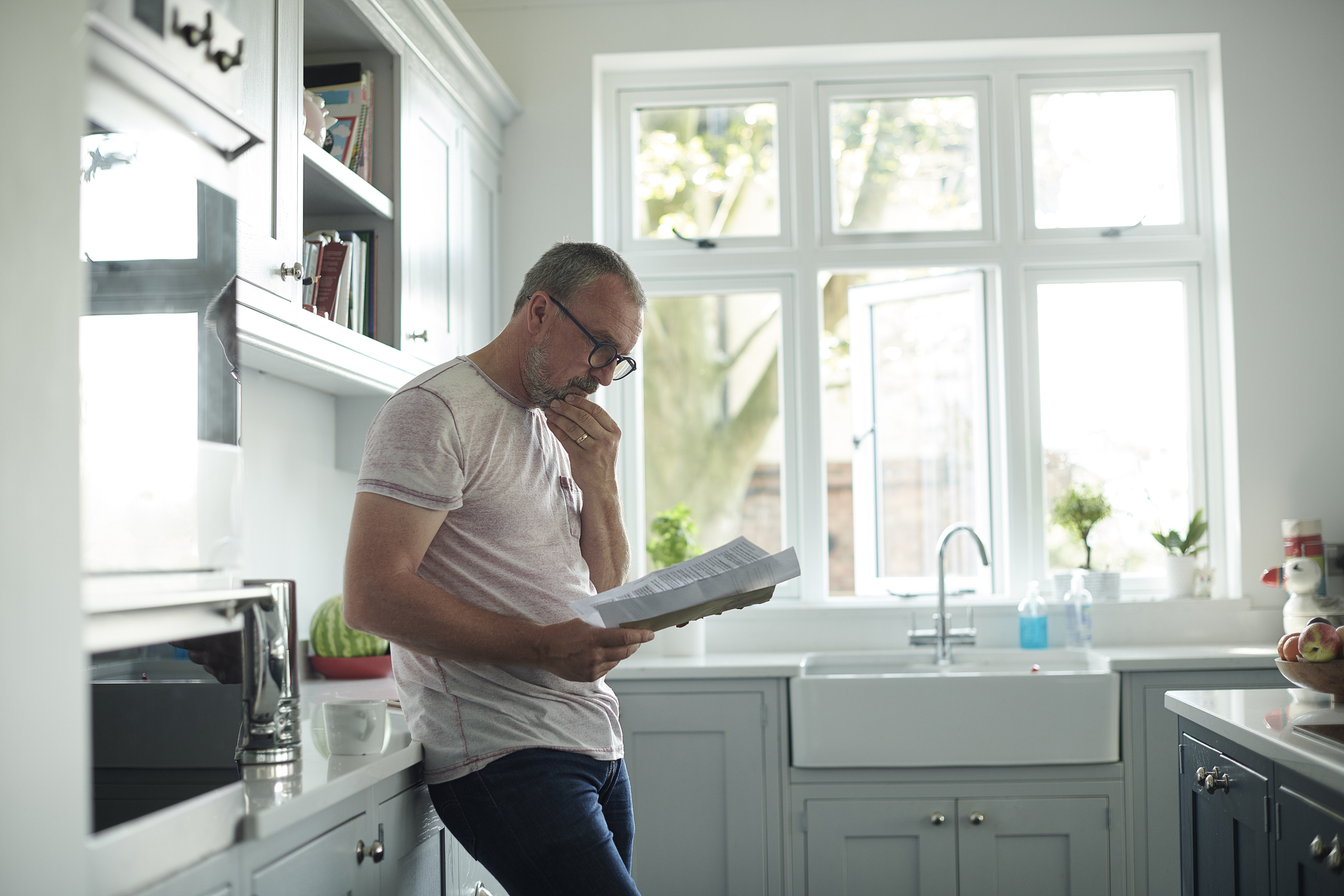 A person standing in a bright, modern kitchen, leaning against the counter while reading a document. Sunlight comes through large windows above the farmhouse-style sink, illuminating the light grey cabinets and countertops.