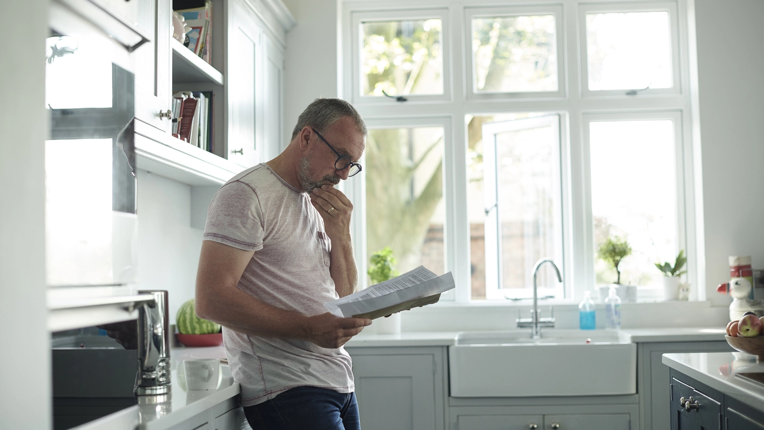 A person standing in a bright, modern kitchen, leaning against the counter while reading a document. Sunlight comes through large windows above the farmhouse-style sink, illuminating the light grey cabinets and countertops. A person standing in a bright, modern kitchen, leaning against the counter while reading a document. Sunlight comes through large windows above the farmhouse-style sink, illuminating the light grey cabinets and countertops.