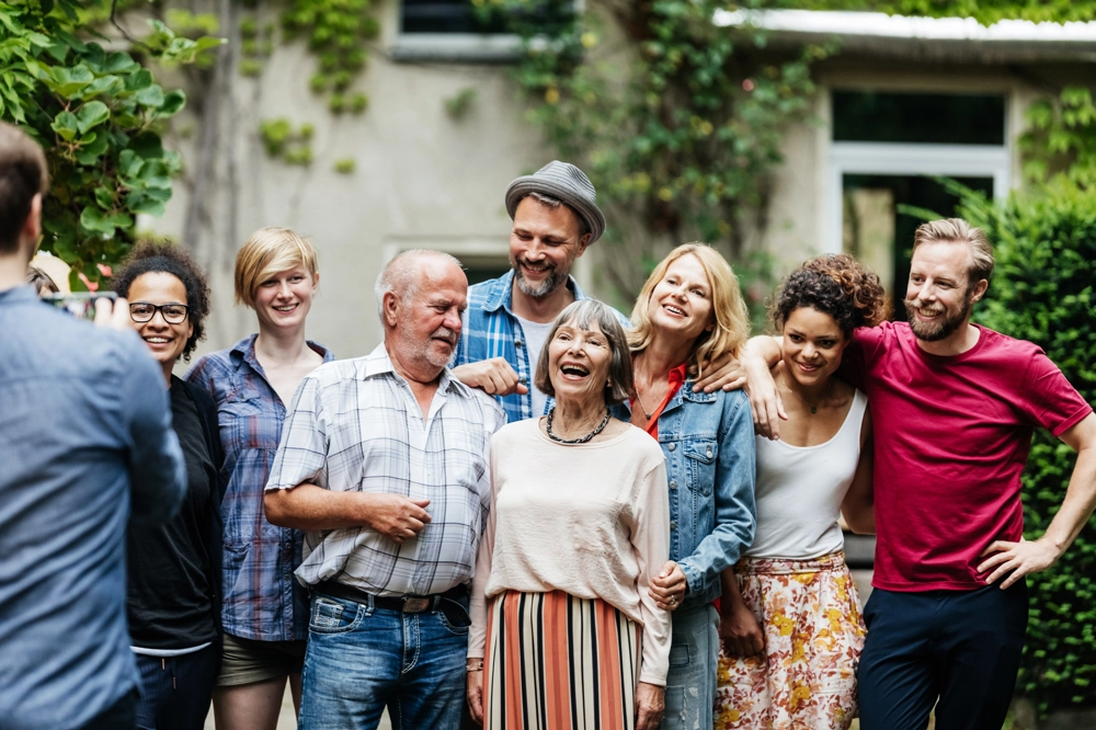 A group of people gathered outside in a leafy back garden, standing close together for a photo. They’re dressed casually, with some resting their arms on each other’s shoulders. The background shows part of a house with windows. A group of people gathered outside in a leafy back garden, standing close together for a photo. They’re dressed casually, with some resting their arms on each other’s shoulders. The background shows part of a house with windows.