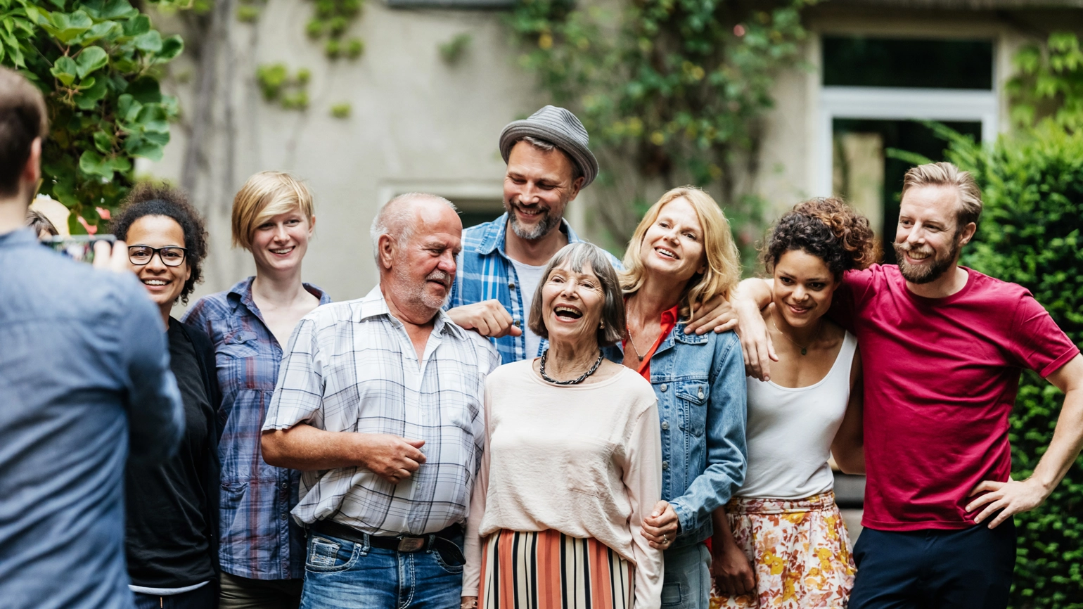 A group of people gathered outside in a leafy back garden, standing close together for a photo. They’re dressed casually, with some resting their arms on each other’s shoulders. The background shows part of a house with windows. A group of people gathered outside in a leafy back garden, standing close together for a photo. They’re dressed casually, with some resting their arms on each other’s shoulders. The background shows part of a house with windows.