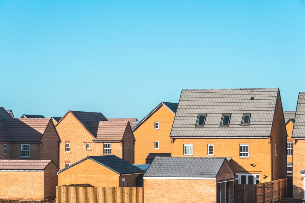 Modern new‑build family houses built in yellow brick with dark pitched tiled roofs under a clear blue daytime sky. Modern new‑build family houses built in yellow brick with dark pitched tiled roofs under a clear blue daytime sky.