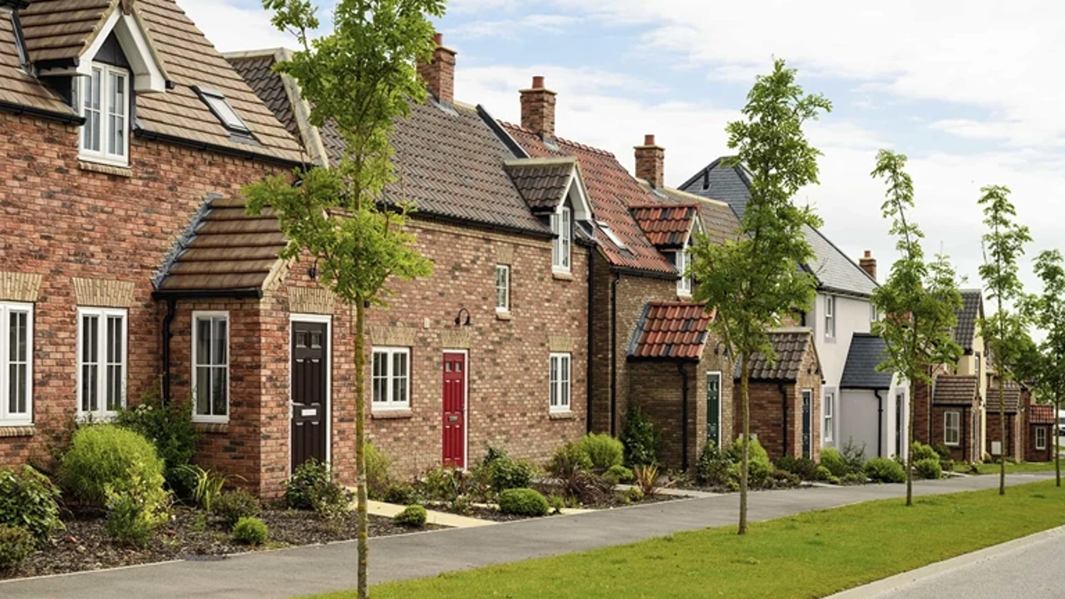 Street view of a suburban housing development; a row of traditional-style brick homes with pitched roofs and varied door colours. Each home has a landscaped front garden, and young trees line a pedestrian pathway. Street view of a suburban housing development; a row of traditional-style brick homes with pitched roofs and varied door colours. Each home has a landscaped front garden, and young trees line a pedestrian pathway.