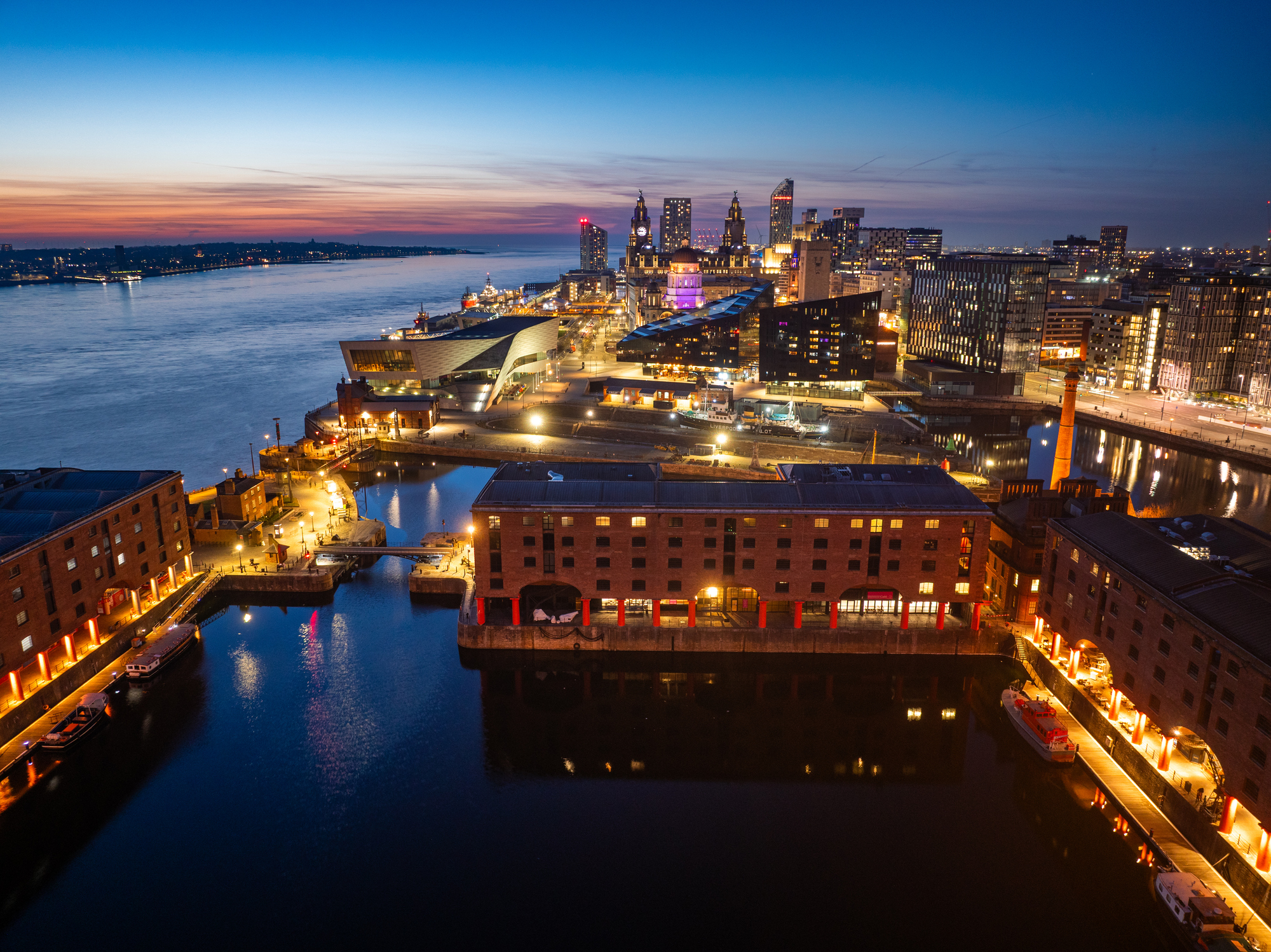 Aerial view of UK city waterfront at dusk, showing illuminated historic dock buildings, modern architecture, and the River