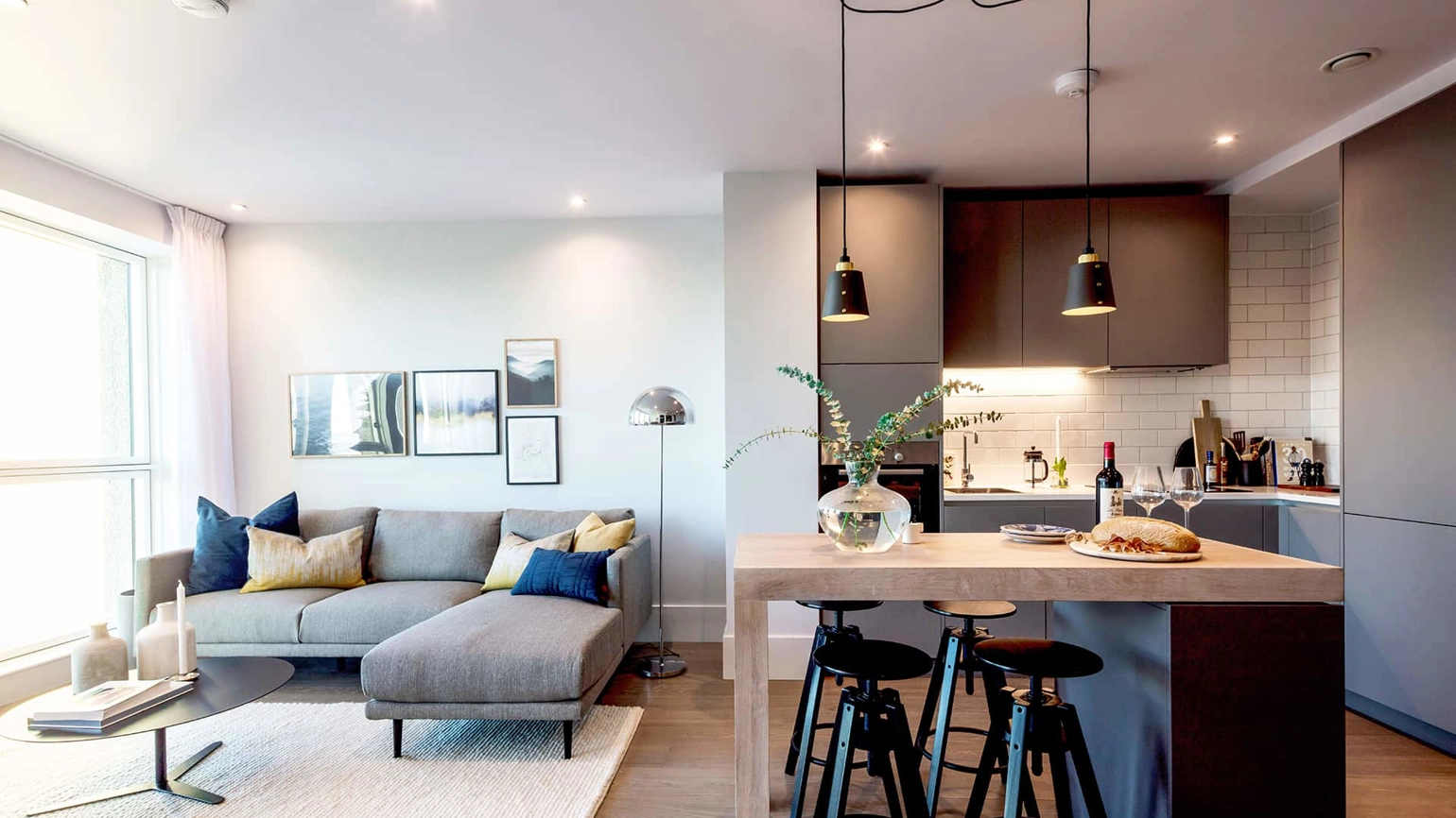 Modern open-plan living space with a light grey sectional sofa and framed artwork on the wall. The space transitions into a sleek kitchen with dark grey cabinetry, white subway tile backsplash, and a wooden island with three black bar stools. Black pendant lights hang above the island, which is set with a glass vase, bottle of wine, and loaf of bread. A large window on the left brings in natural light. Modern open-plan living space with a light grey sectional sofa and framed artwork on the wall. The space transitions into a sleek kitchen with dark grey cabinetry, white subway tile backsplash, and a wooden island with three black bar stools. Black pendant lights hang above the island, which is set with a glass vase, bottle of wine, and loaf of bread. A large window on the left brings in natural light.