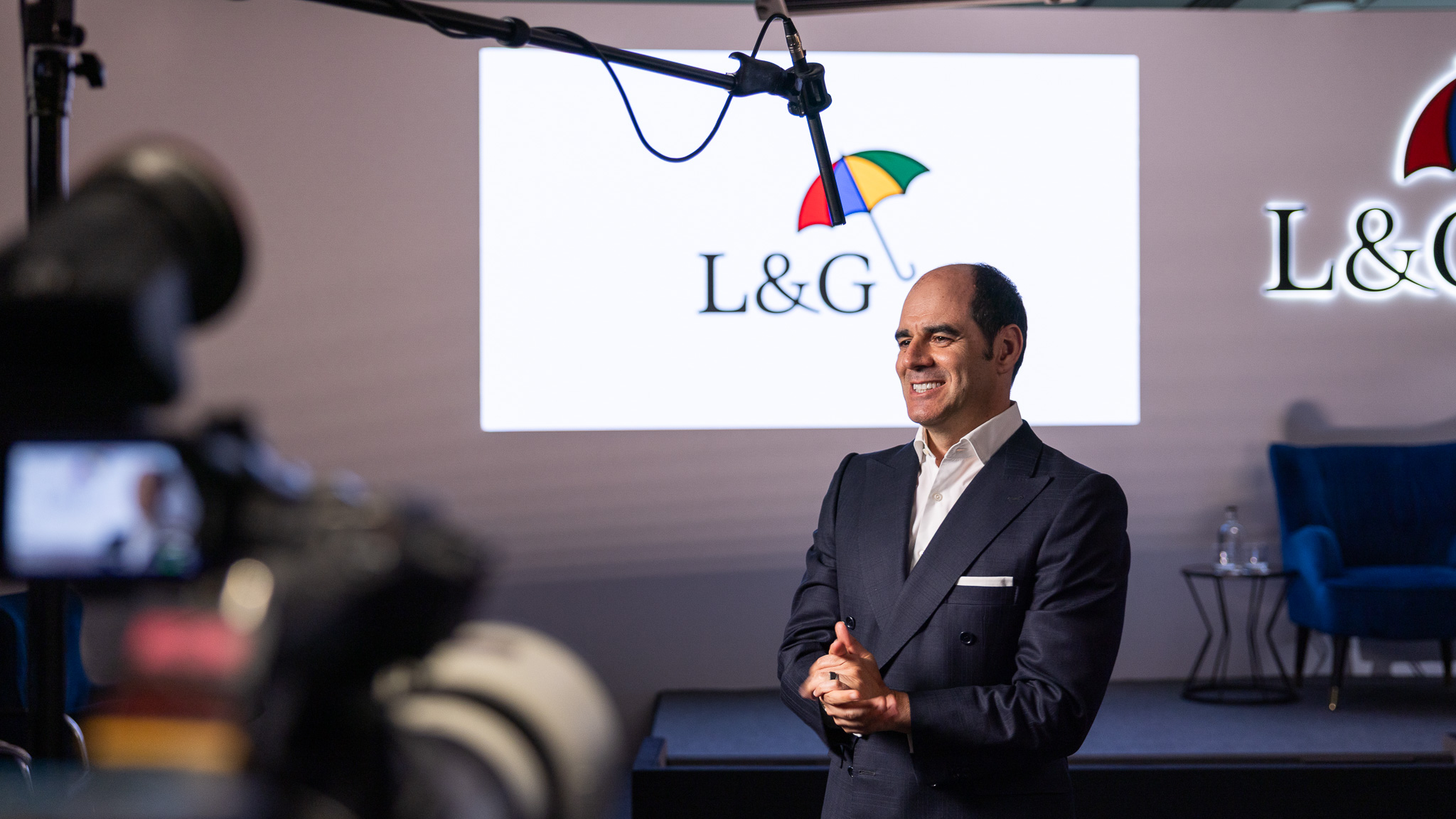  António Simões is standing in front of an empty stage, with the L&G logo projected on the wall. He is wearing a dark suit and white shirt, with his hands clasped in front of him. A microphone boom is positioned above him, and there are cameras and lighting equipment visible in the foreground.