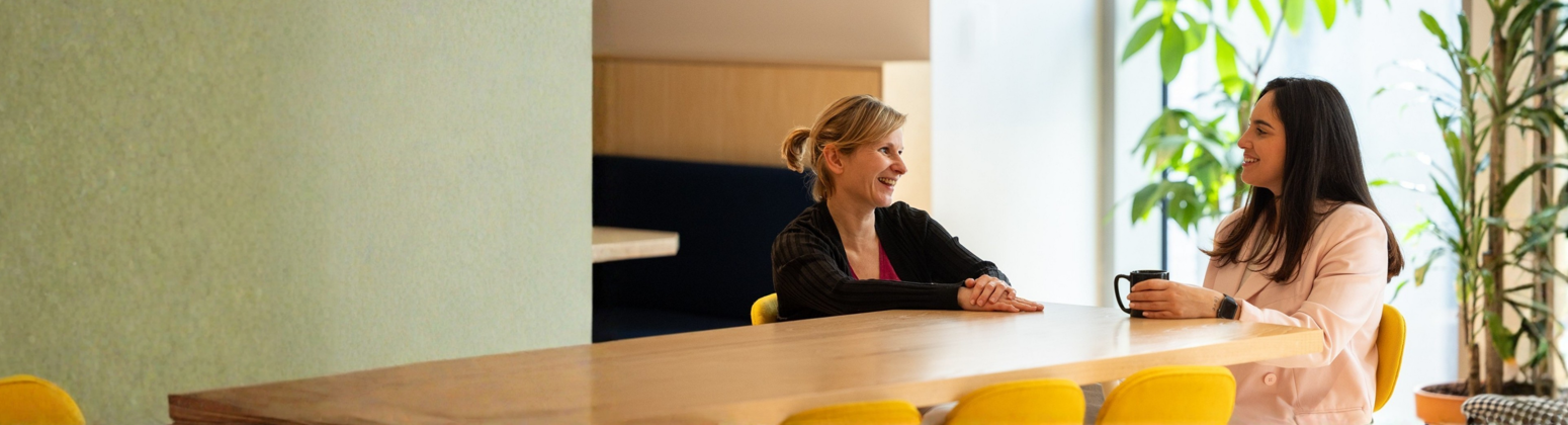 Two colleagues seated at a wooden table with yellow chairs in a bright, plant-filled room with large windows. The person on the left wears a dark jacket, while the one on the right wears a light blazer and holds a black mug. They appear to be engaged in a conversation. Two colleagues seated at a wooden table with yellow chairs in a bright, plant-filled room with large windows. The person on the left wears a dark jacket, while the one on the right wears a light blazer and holds a black mug. They appear to be engaged in a conversation.