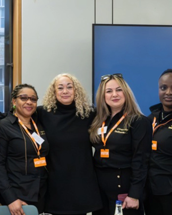 Six people stand together indoors, all wearing black clothing and orange visitor badges. Six people stand together indoors, all wearing black clothing and orange visitor badges.