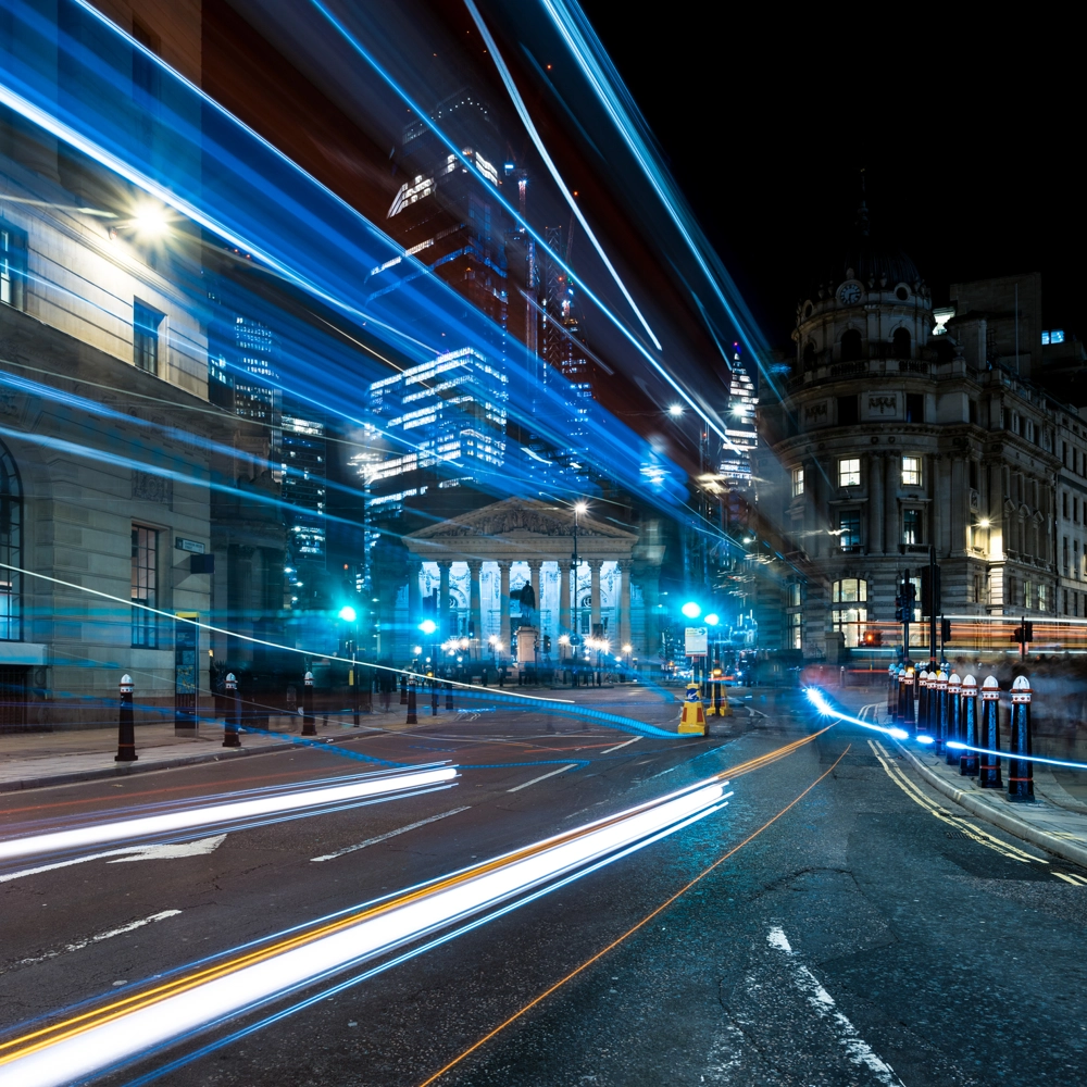 Night view of the Bank Junction in London, UK, with illuminated buildings and skyscrapers in the background. Bright streetlights create starburst effects, and light trails from passing vehicles flow across the foreground. Night view of the Bank Junction in London, UK, with illuminated buildings and skyscrapers in the background. Bright streetlights create starburst effects, and light trails from passing vehicles flow across the foreground.