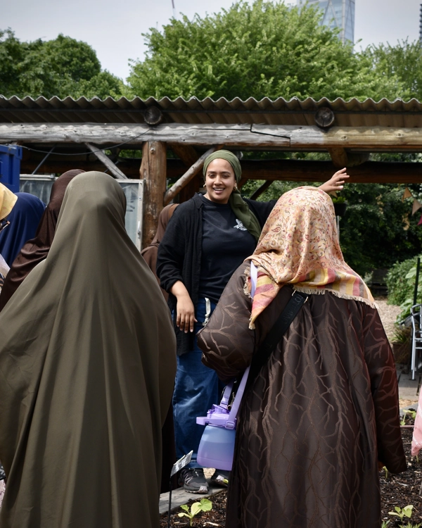 A group of women in hijabs listen to a speaker outdoors, with trees and a wooden structure in the background. A group of women in hijabs listen to a speaker outdoors, with trees and a wooden structure in the background.