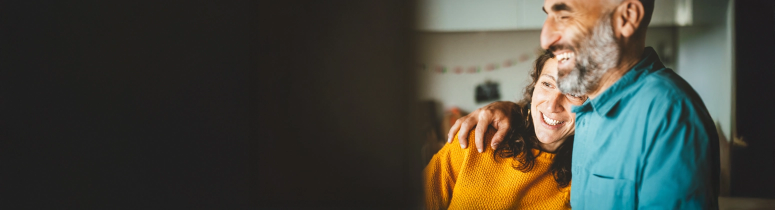 A person in a teal shirt with an arm around another person wearing a yellow sweater inside a softly lit kitchen. A person in a teal shirt with an arm around another person wearing a yellow sweater inside a softly lit kitchen.