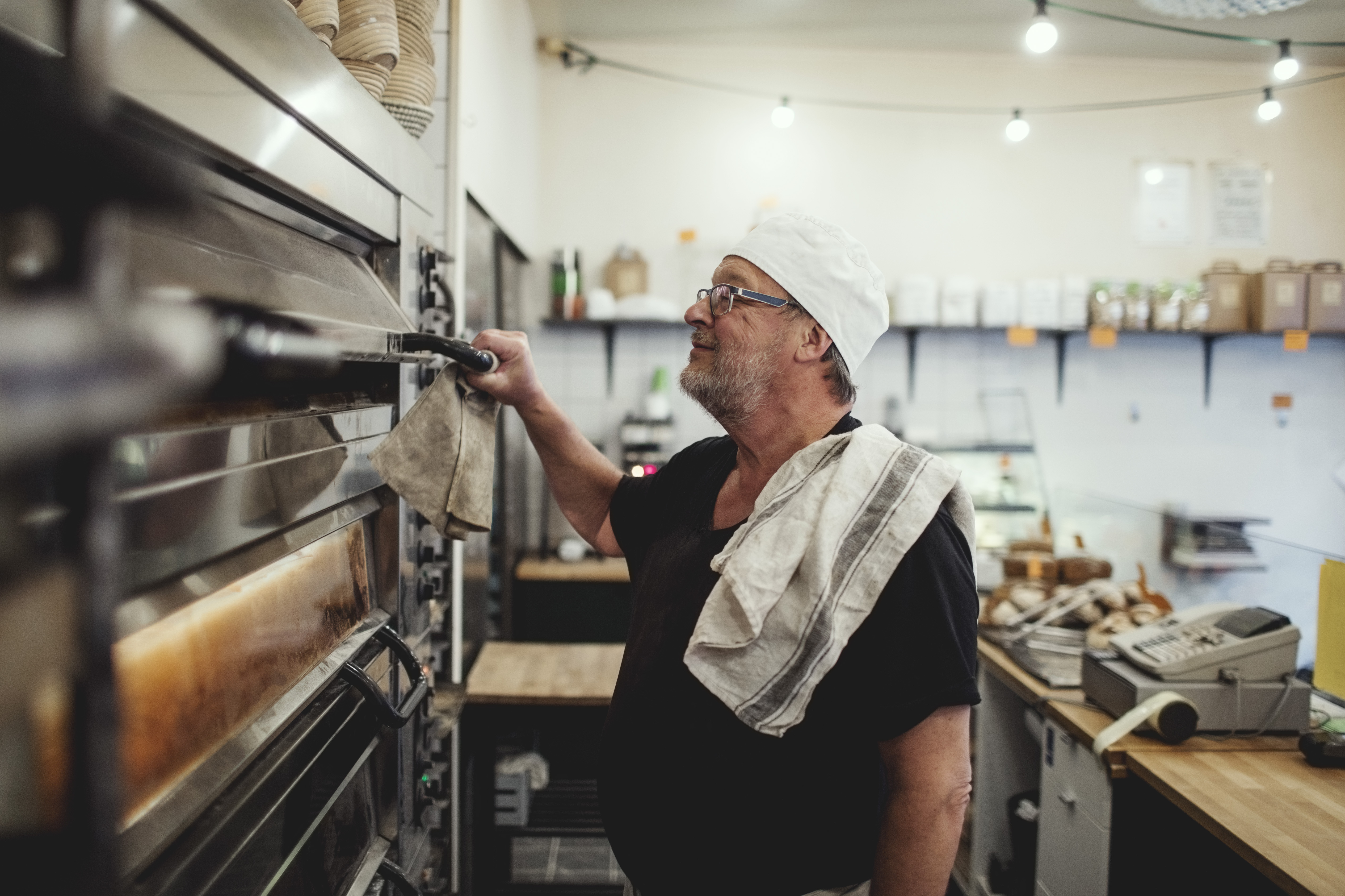 A senior baker stands by a oven at a small bakery.
