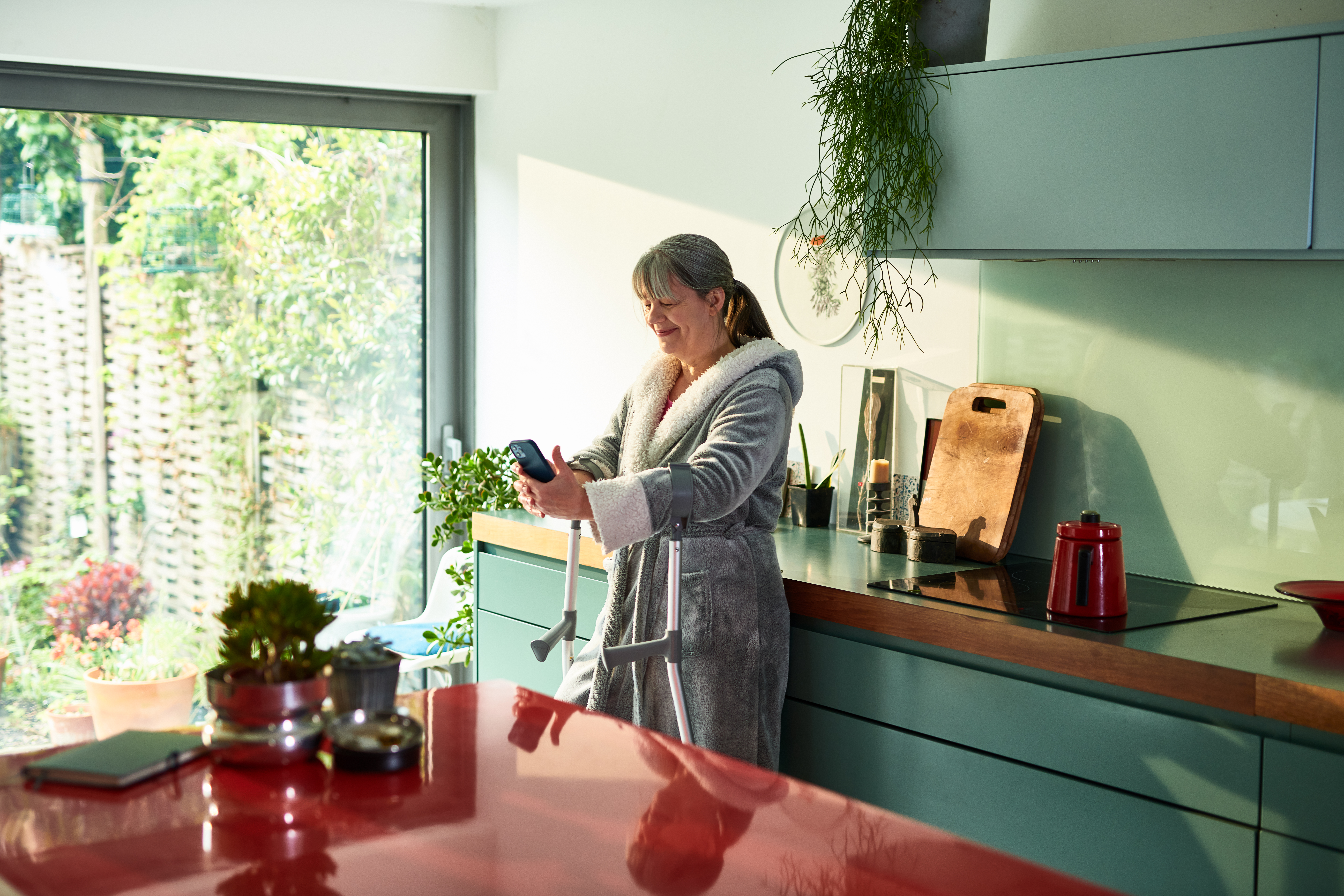 A smiling woman standing in a kitchen focusing on her smart phone.