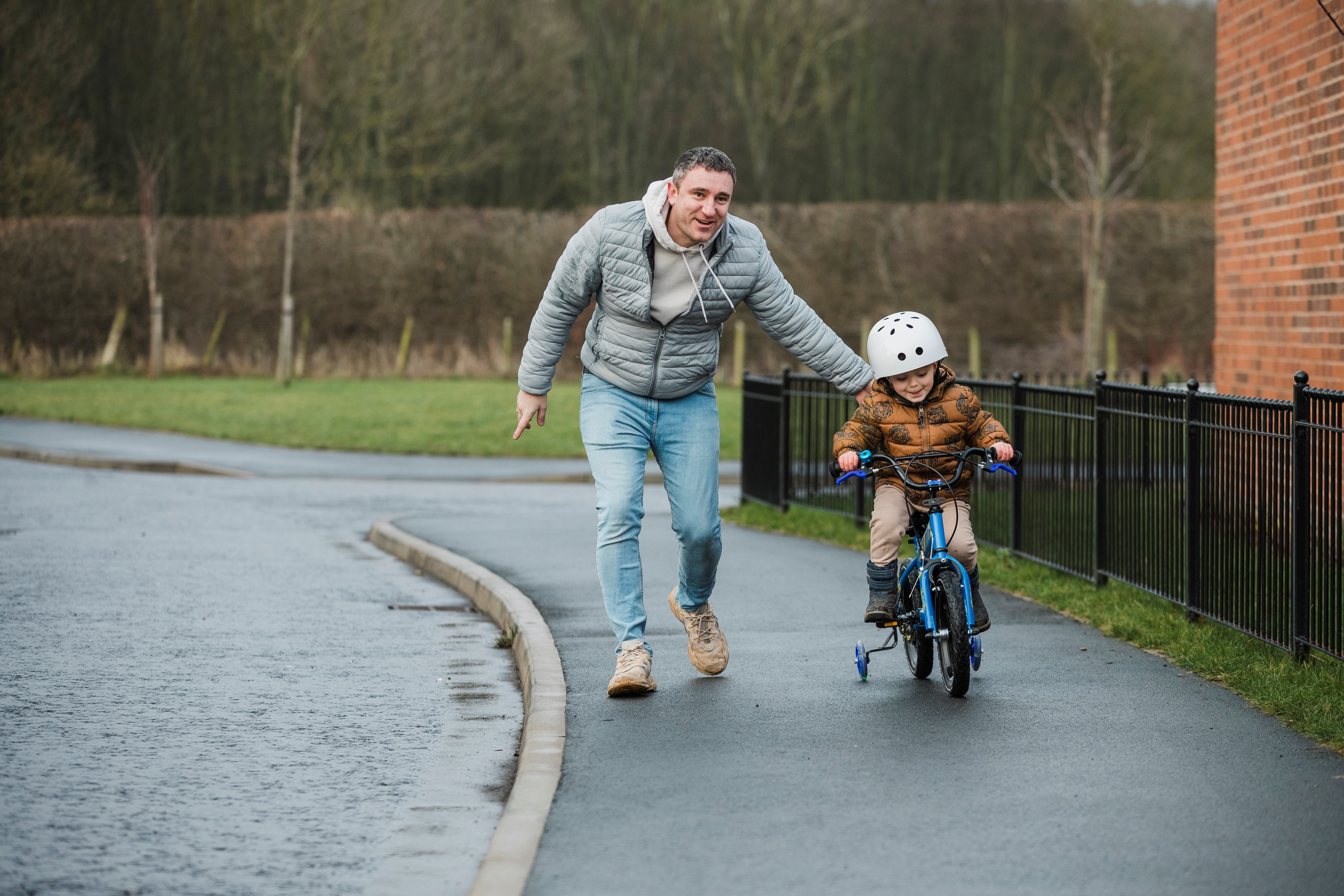 A father rests a reassuring hand on his young son's back as he learns to ride his bike on the footpath outside of his home in the North East of England. The young boy is smiling as he controls the bike on the footpath.