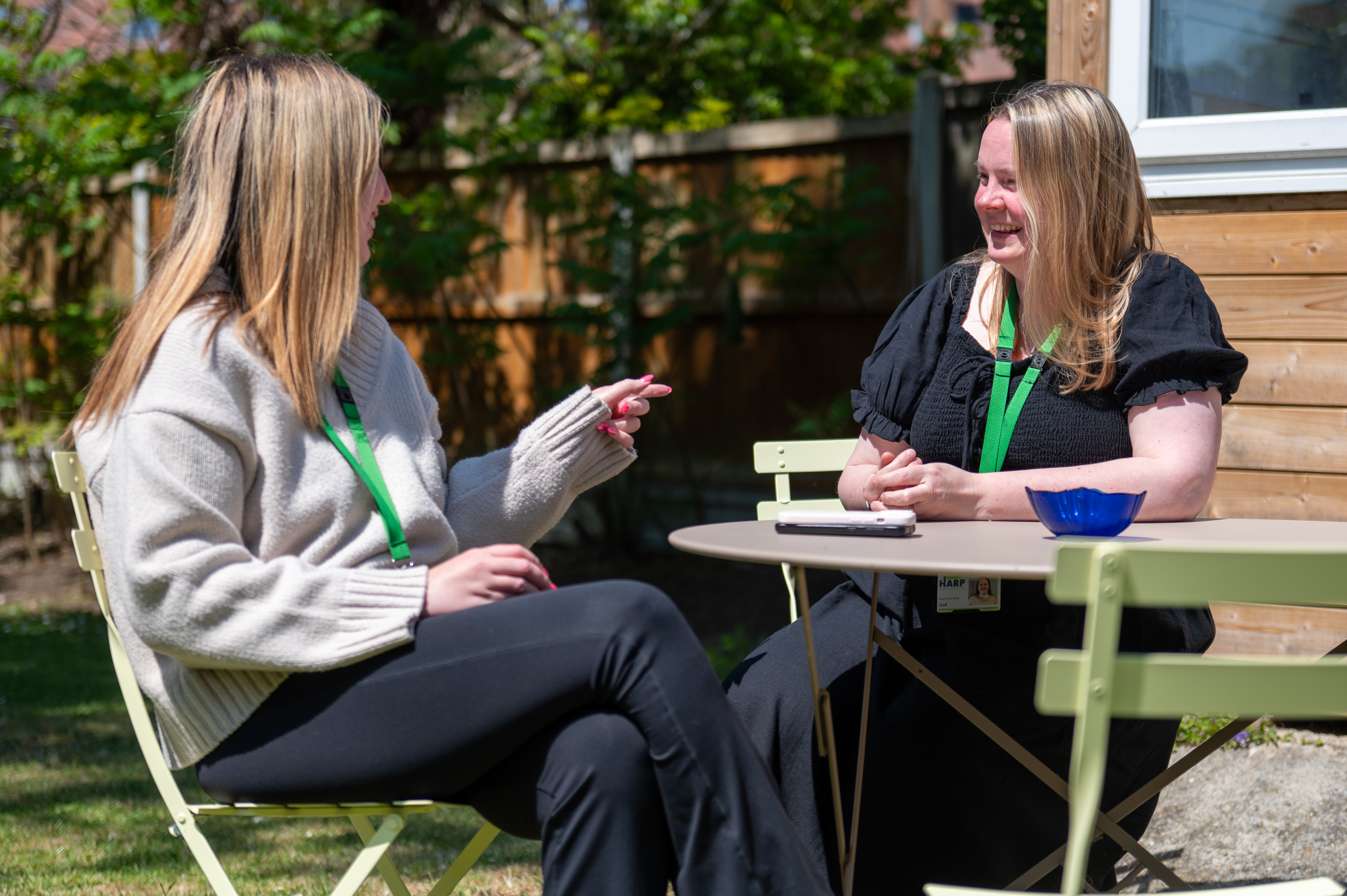 Two people with blurred faces sit at a round outdoor table talking. Both wear green lanyards; a blue bowl and smartphone are on the table. Greenery and a wooden fence are in the background.