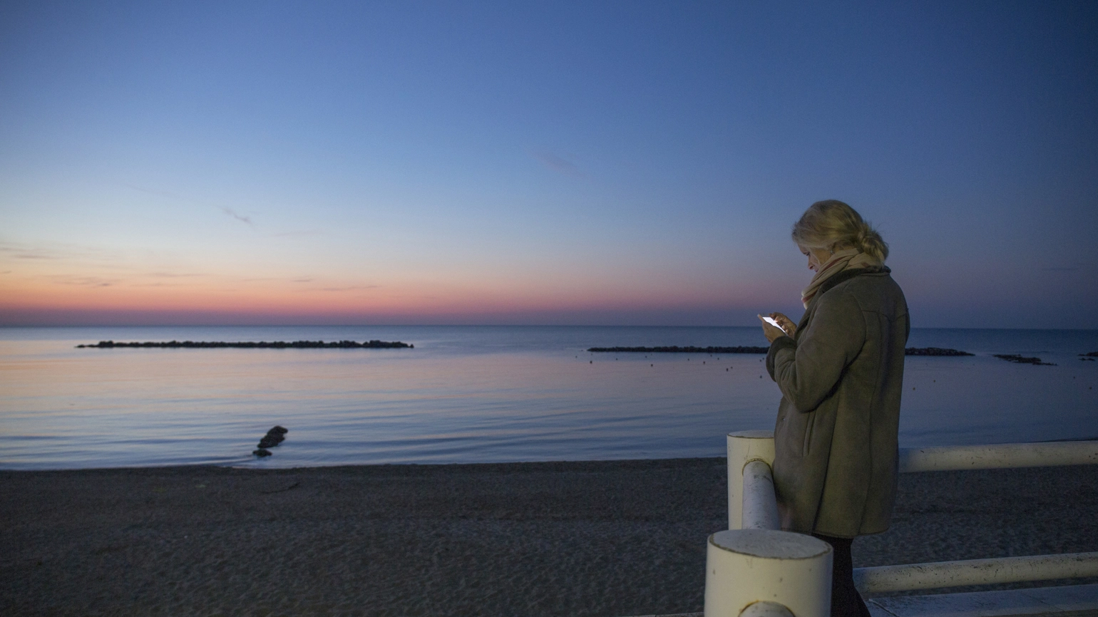 A person wearing a winter coat standing on a seaside promenade at dusk, illuminated by the light of their phone while facing a calm ocean and a sky that transitions from deep blue to soft pink near the horizon. A person wearing a winter coat standing on a seaside promenade at dusk, illuminated by the light of their phone while facing a calm ocean and a sky that transitions from deep blue to soft pink near the horizon.