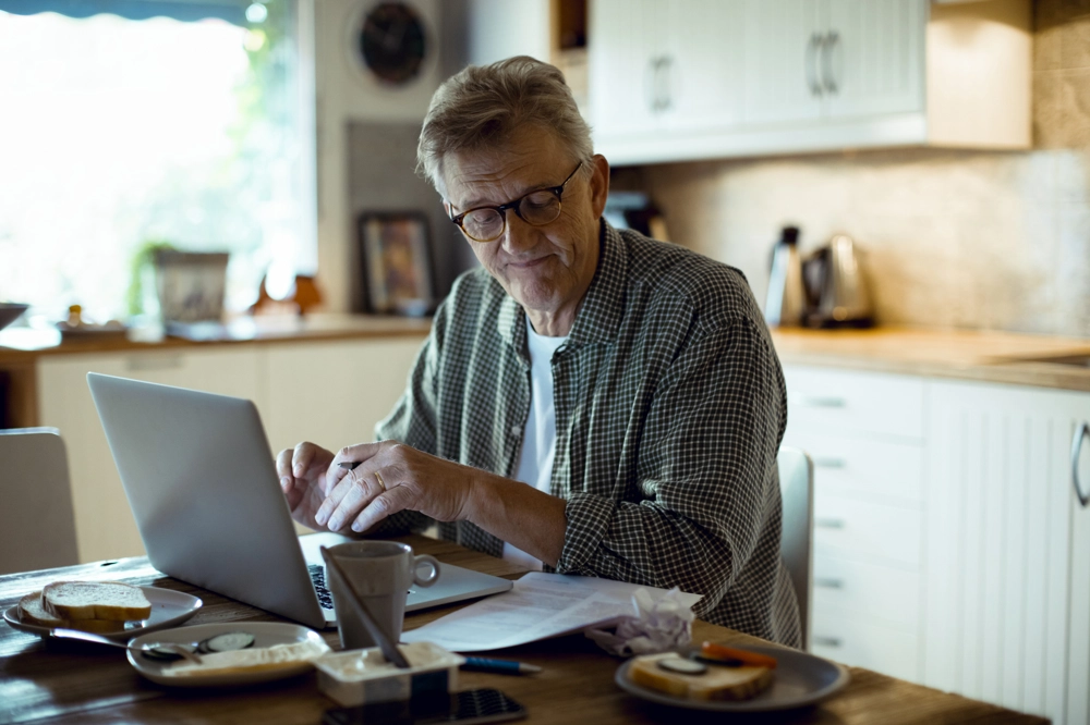 Person sitting at a kitchen table using a laptop, with papers and a coffee cup spread out on the table. The background shows a bright, homey kitchen with white cabinets and a kettle on the counter. Person sitting at a kitchen table using a laptop, with papers and a coffee cup spread out on the table. The background shows a bright, homey kitchen with white cabinets and a kettle on the counter.