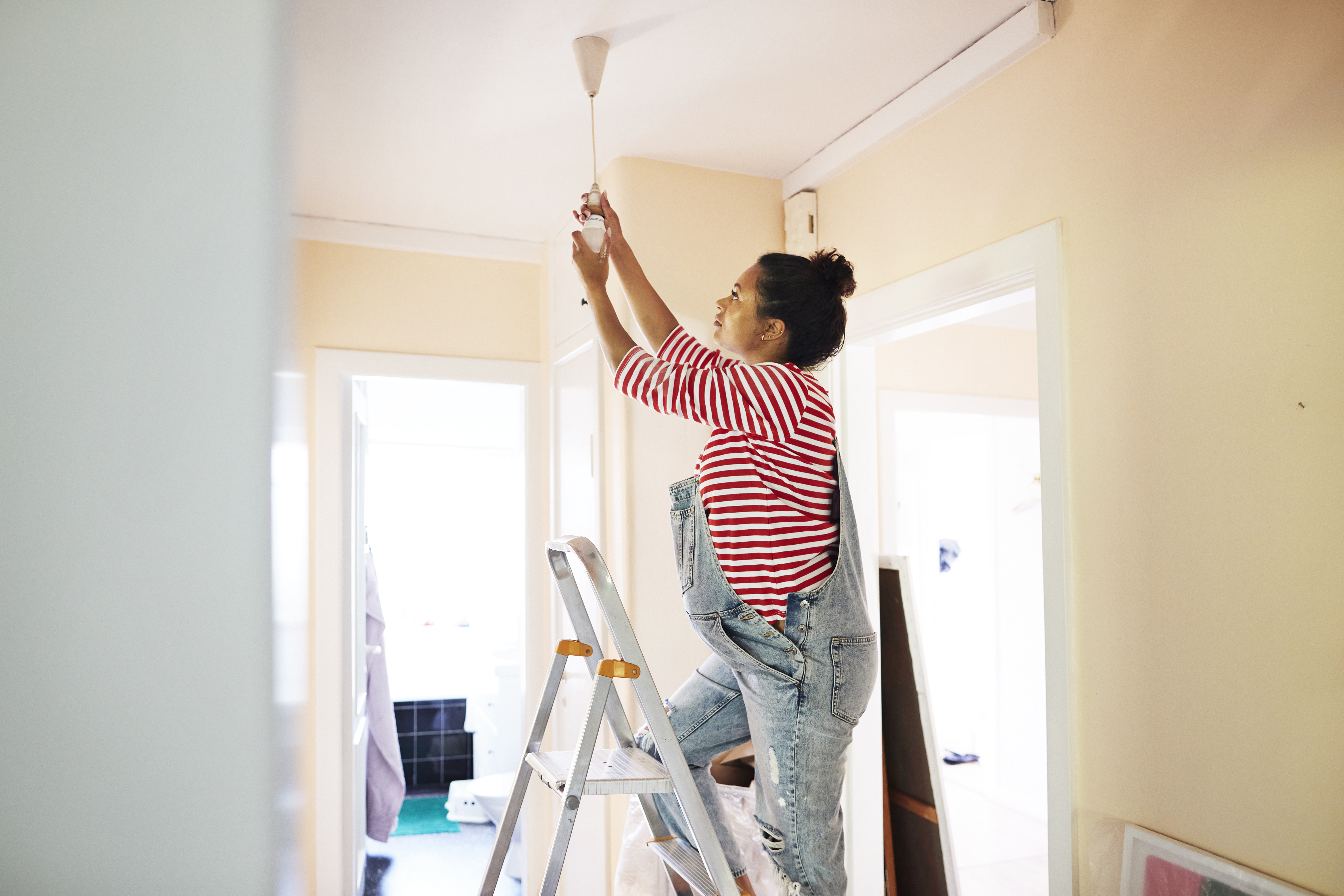A pregnant woman changing a light bulb on a corridor ceiling while standing on ladder.