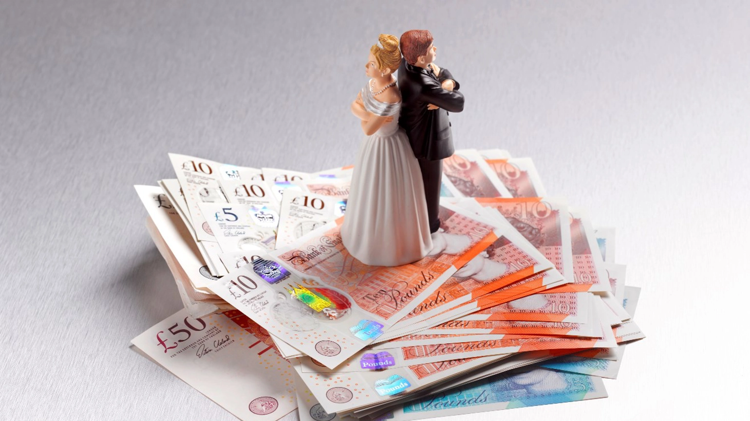 Wedding cake topper of a bride and groom standing back-to-back with arms crossed, placed on a stack of banknotes. Wedding cake topper of a bride and groom standing back-to-back with arms crossed, placed on a stack of banknotes.