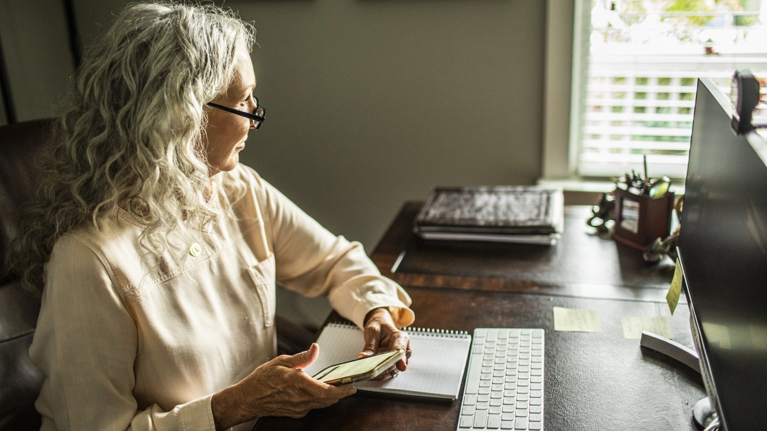 Senior woman holding a smartphone while looking out a window in her home office. Senior woman holding a smartphone while looking out a window in her home office.
