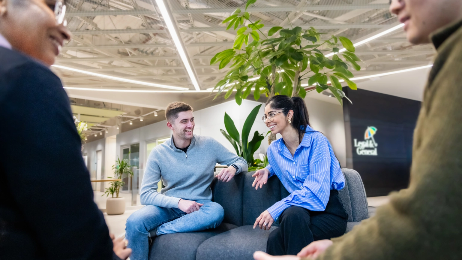 Four people seated on a dark grey circular sofa in Legal & General’s London office. Plants are arranged in the centre of the seating area, behind the people. Four people seated on a dark grey circular sofa in Legal & General’s London office. Plants are arranged in the centre of the seating area, behind the people.