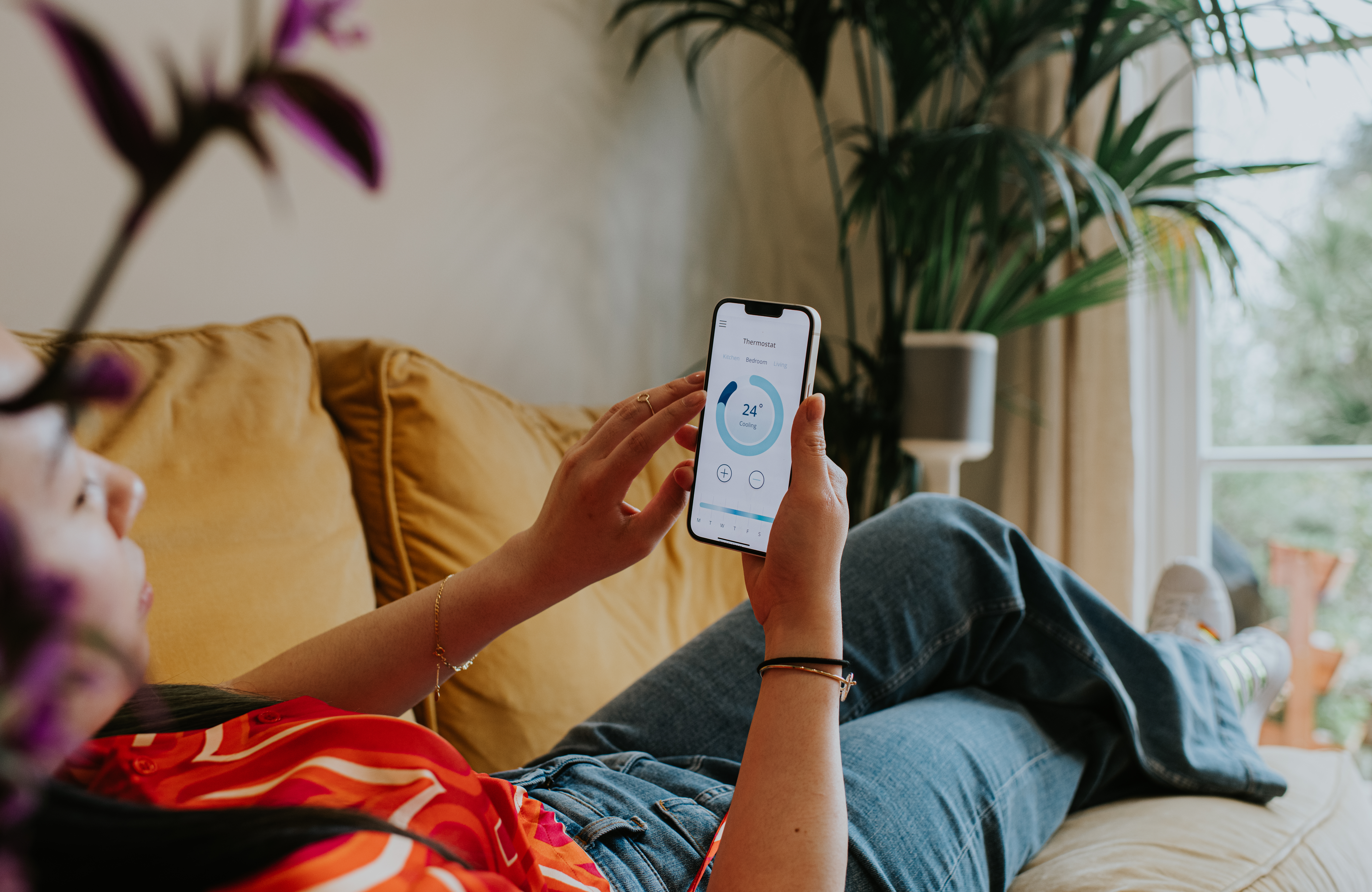 A woman with red shirt lies down on a sofa and uses her smartphone to check her home thermostat